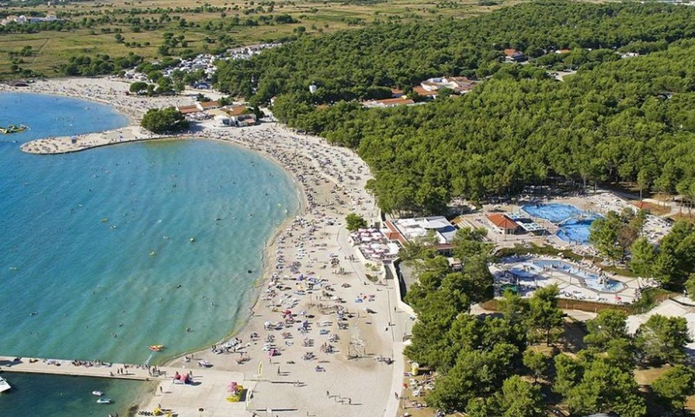 Zračni pogled na plažu Zaton u blizini Zadra s pješčanom obalom, kristalno čistim morem i okolnom borovom šumom koja nudi rekreacijske sadržaje.