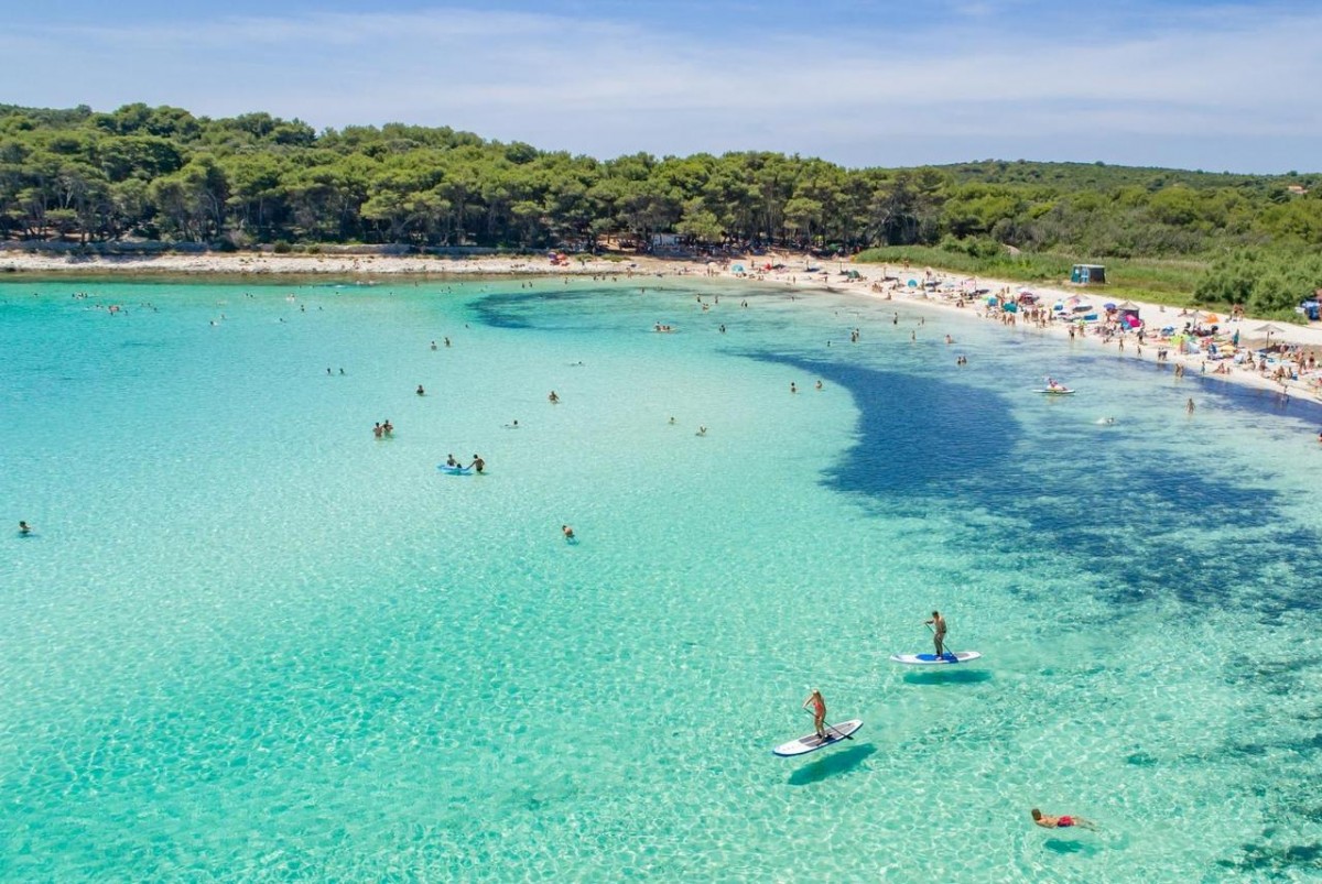 Prekrasan pogled na plažu Sakarun na Dugom otoku s tirkizno čistim morem, bijelim pijeskom i ljudima koji uživaju u kupanju i veslanju.