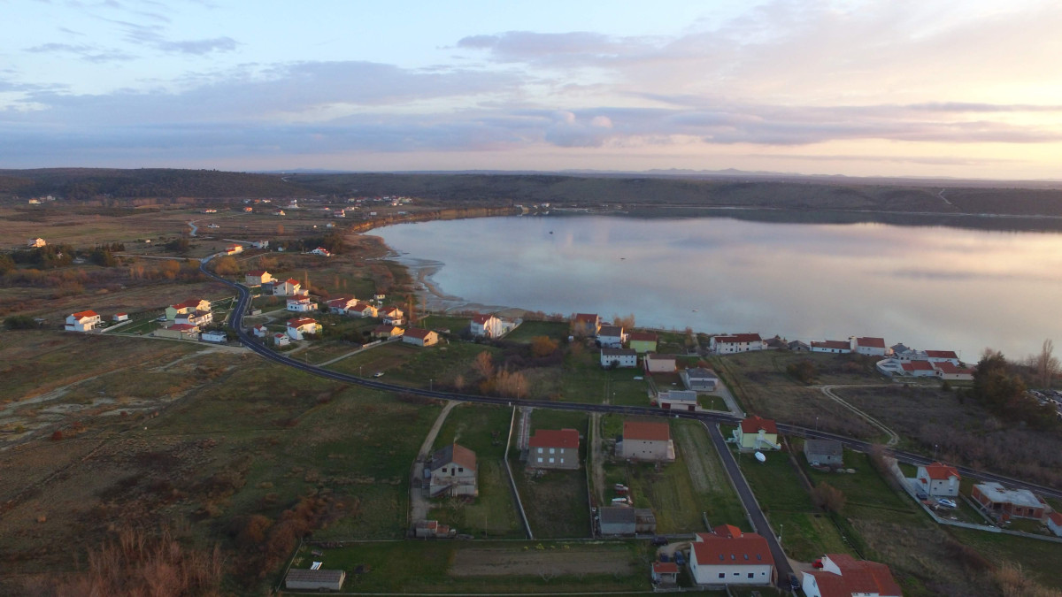 Zračni pogled na plažu Ljubač, mirnu i plitku uvalu okruženu kućama, zelenim poljima i slikovitim krajolicima pod blagim zalaskom sunca.